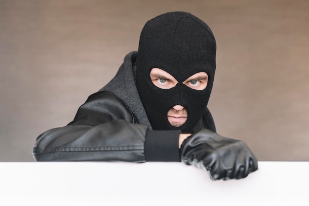 Foto un ladro malvagio in una maschera guarda fuori da dietro un bandito malvagio da tavola bianca con una maschera sul viso