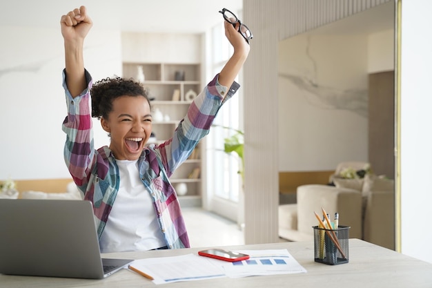 Photo exam is passed happy teenage girl is sitting in front of laptop at workplace and raising hands up