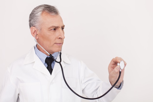 Examining a copy space. Senior grey hair doctor in uniform looking away and holding his stethoscope outstretched while standing isolated on white