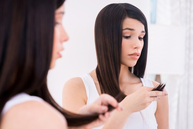 Examining her damaged hair. Frustrated young woman looking at her hair and expressing negativity while standing against mirror