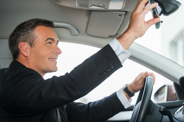 Examining his new car. Side view of cheerful mature man in formalwear adjusting mirror while sitting in his car
