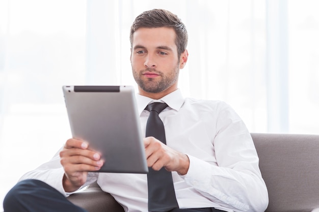 Examining his new tablet. Confident young businessman in shirt and tie working on digital tablet while sitting at the chair