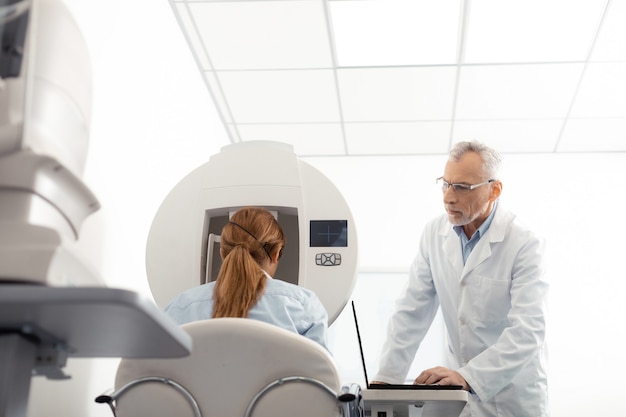 Examining woman attentively. Grey-haired eye specialist wearing glasses examining woman attentively