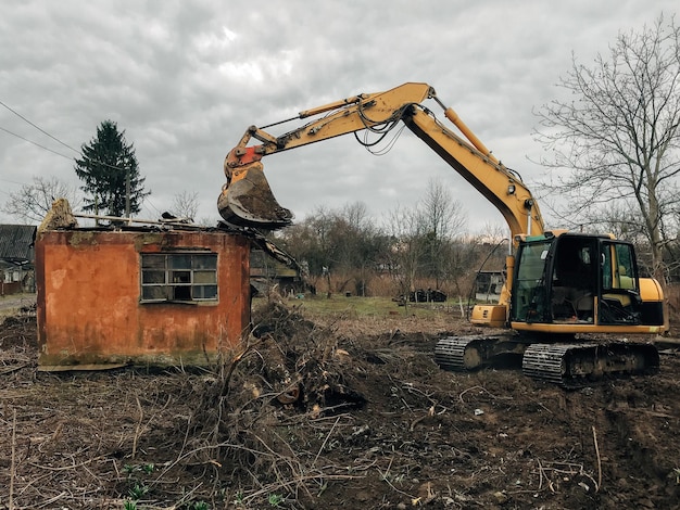 Excavator destroying brick house on land in countryside\
bulldozer clearing land from old bricks and concrete from walls\
with dirt and trash backhoe machinery ruining house