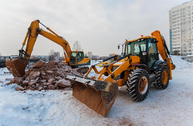 An excavator digs the ground at a construction site in winter against the background of a new house