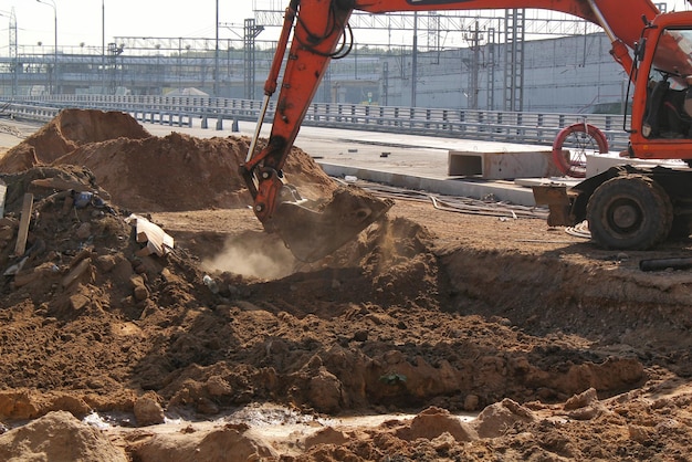 도시로 가는 새로운 도로 건설 현장에서 굴삭기 기계가 작동합니다. 오렌지 굴착기는 모래를 파다