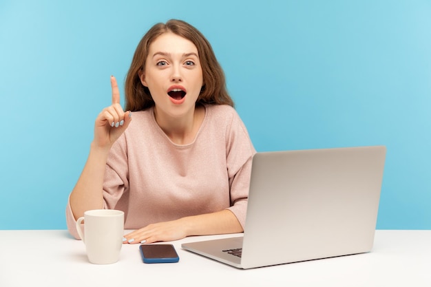 Excellent business solution Excited young businesswoman looking surprised by genius idea raising finger up inspired by creative thought at workplace indoor studio shot isolated on blue background