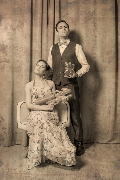 Foto excentriek huwelijk portret. de vrouw, gezeten in een elegante gebloemde jurk, draagt een speelgoedwapen. de man, staand, gekleed in een elegant pak en met een speelgoedkatje. ernstige en plechtige uitdrukking.