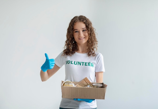 Excited activist volunteer holding box with donations and showing thumb up smiling at camera over light background