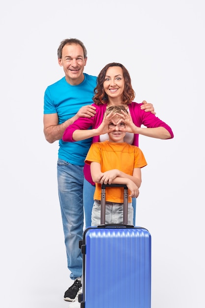 Photo excited adult mother smiling and gesturing glasses on face of boy with suitcase while traveling with husband and son against white background