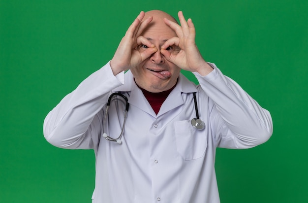 Excited adult slavic man in doctor uniform with stethoscope stucking out his tongue and through fingers 