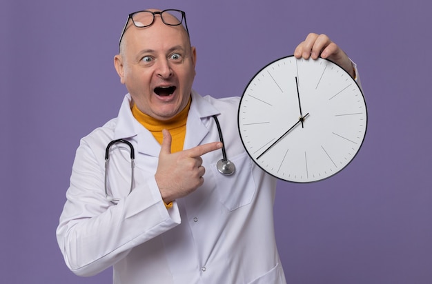 Uomo slavo adulto eccitato con occhiali ottici in uniforme da medico con stetoscopio che tiene e punta all'orologio