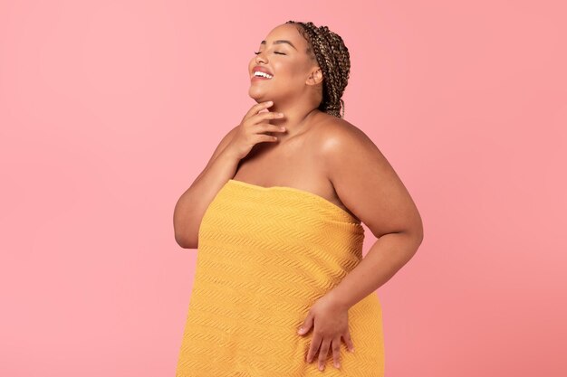 Excited african american body positive lady wrapped in towel touching her soft skin on neck enjoying her young beauty