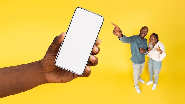 Excited African American Couple Pointing At Large Smartphone Yellow Background