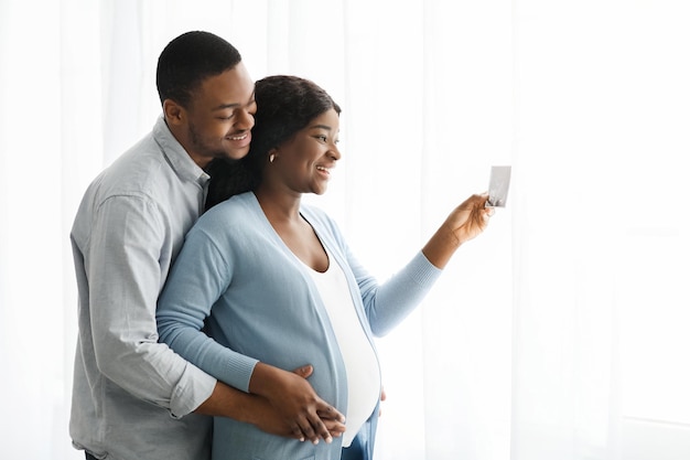 Famiglia in attesa afroamericana eccitata che guarda l'immagine ad ultrasuoni del loro bambino. felice uomo di colore che abbraccia sua moglie incinta con ecografia del bambino, in piedi vicino alla finestra a casa, vista laterale, spazio copia