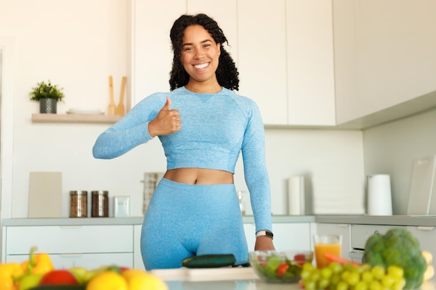 Excited african american fitness woman in sportswear showing thumb up standing in kitchen after domestic workout