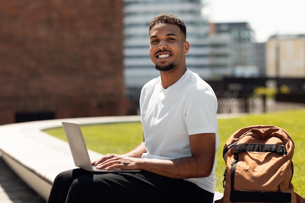 흥분한 아프리카계 미국인 남성 프리랜서가 노트북 컴퓨터 작업을 하고 야외에 앉아 웃고 있습니다.