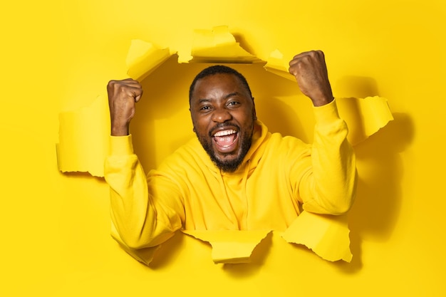 Excited african american man raising clenched fists celebrating success and smiling posing in ripped yellow hole