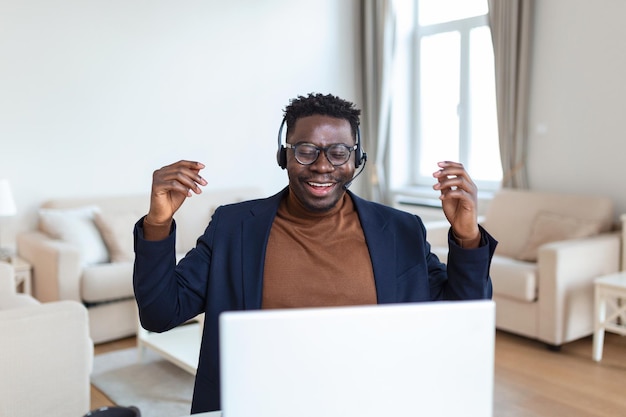 Возбужденный афроамериканец в наушниках читает хорошие новости по электронной почте, получает новую работу, продвигается по службе, использует ноутбук, смотрит на экран и кричит от радости, показывает жест "да", празднует