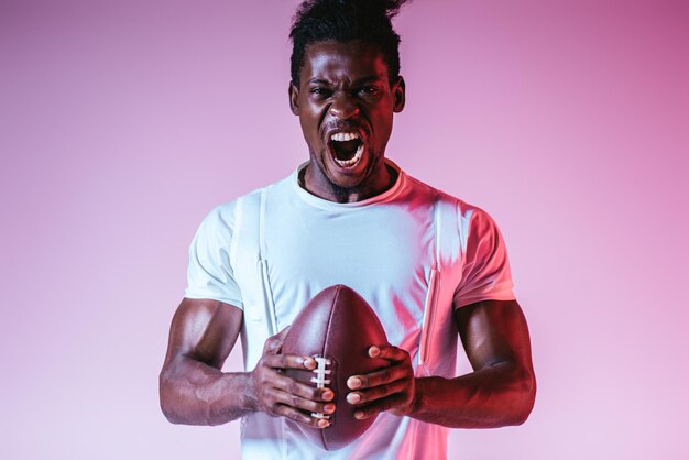 Excited african american sportsman yelling at camera while holding rugby ball on purple background
