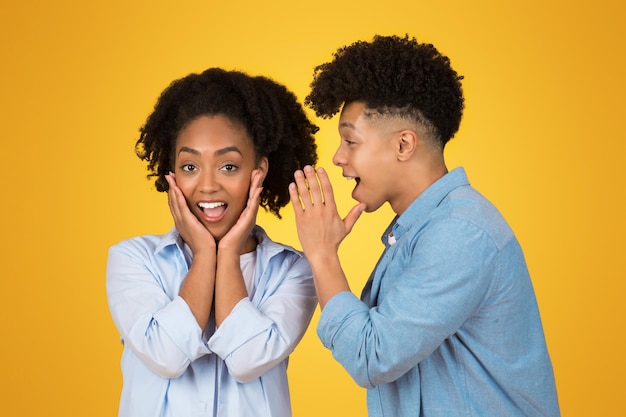 An excited african american woman with hands on her cheeks in surprise while a man whispers