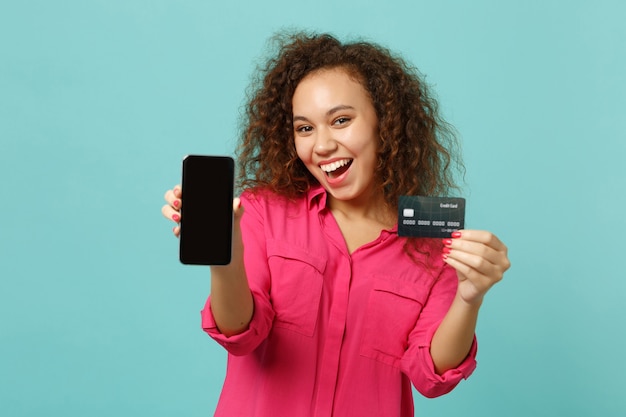 La ragazza africana emozionante in vestiti casuali tiene il telefono cellulare con lo schermo vuoto in bianco, carta bancaria di credito isolata su fondo blu del turchese. concetto di stile di vita di emozioni sincere della gente. mock up copia spazio.