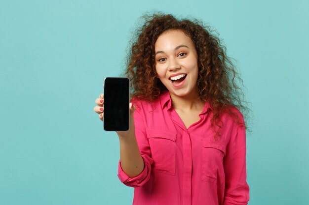 Excited african girl in casual clothes hold mobile phone with blank empty screen isolated on blue turquoise wall background in studio. People sincere emotions, lifestyle concept. Mock up copy space.