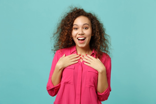 Excited african girl in casual clothes keeping mouth open, holding hands on chest isolated on blue turquoise wall background in studio. People sincere emotions, lifestyle concept. Mock up copy space.