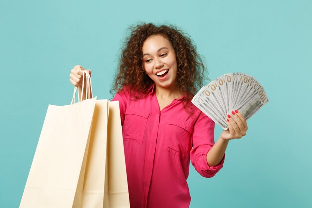 Excited african girl hold package bag with purchases after shopping, fan of money in dollar banknotes, cash money isolated on blue turquoise background. People lifestyle concept. Mock up copy space.