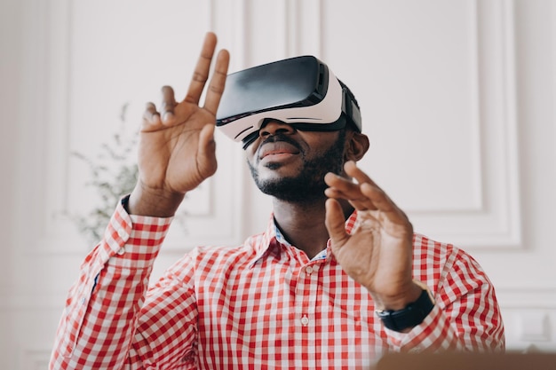 Excited African male in VR glasses explores augmented reality waving hands making touch gestures against home office backdrop