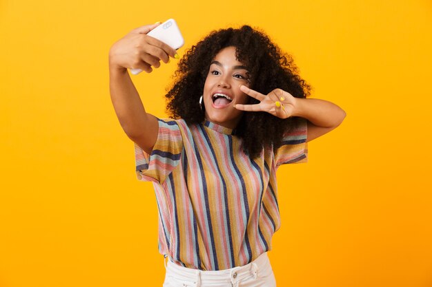 Excited african woman posing isolated over yellow space take a selfie by mobile phone showing peace gesture.