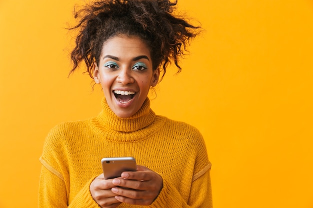 Excited african woman wearing sweater standing isolated, holding mobile phone