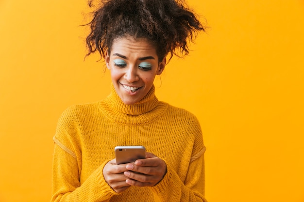 Excited african woman wearing sweater standing isolated, holding mobile phone