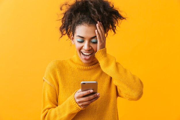 Excited african woman wearing sweater standing isolated, holding mobile phone