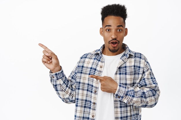 Excited and amazed african american guy, pointing fingers left at sale logo, showing banner, gasp in awe, staring impressed at camera, white background. Copy space