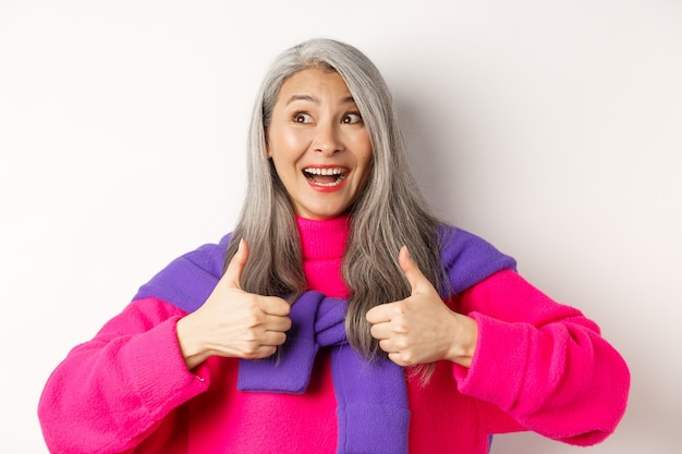 Excited and amazed asian elderly woman showing thumbs-up in approval, smiling happy at upper left corner, praising something awesome, white background