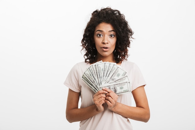 Excited american woman with afro hairstyle holding lots of money dollar currency, isolated over white wall