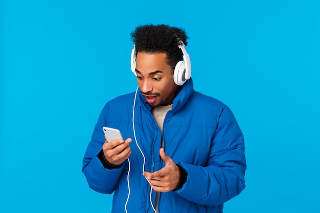 Excited and amused, happy smiling african-american man seeing favorite artist uploaded new song, holding phone press play as listening music in headphones standing winter street, blue wall