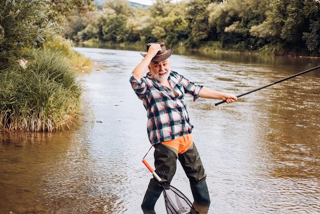 Excited angler senior man fisherman in cowboy hat with fishing rod spinning reel on river old man ca