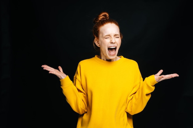 Excited angry young woman in yellow sweater screaming with closed eyes on isolated black background looking at camera Pretty redhead lady model emotionally showing facial expressions copy space