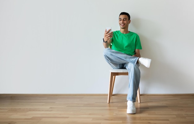 Excited arab guy using cellphone taking selfie or video calling sitting on chair over light wall free space