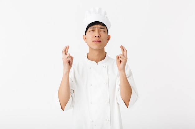 Excited asian chef wearing uniform standing isolated over white wall, holding fingers crossed for good luck