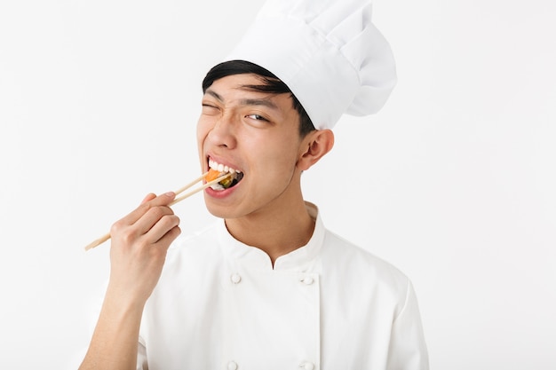 Excited asian chef wearing uniform standing isolated over white wall, preparing sushi