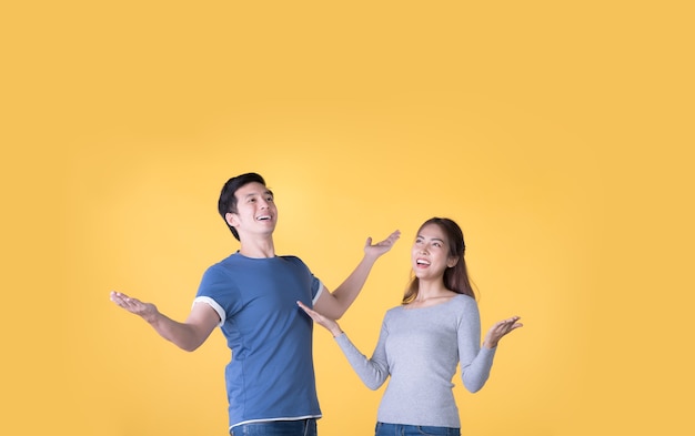 Excited Asian couple in casual clothes presenting with hands open to copy space isolated on yellow background