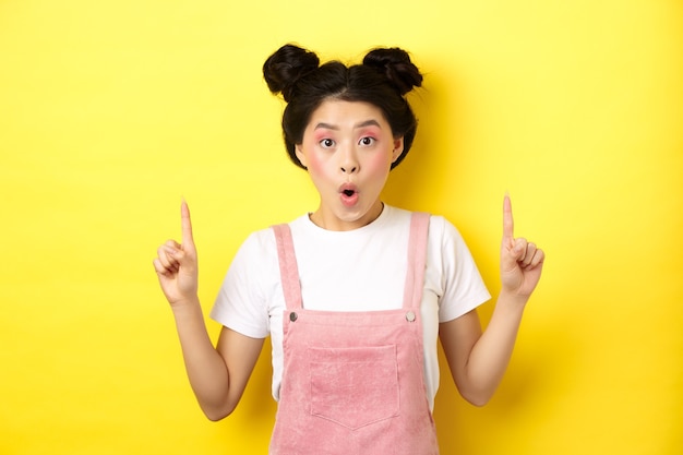Возбужденная азиатская модель с гламурным макияжем, указывая пальцами вверх и изумившись, проверяет промо-предложение, стоя на желтом фоне