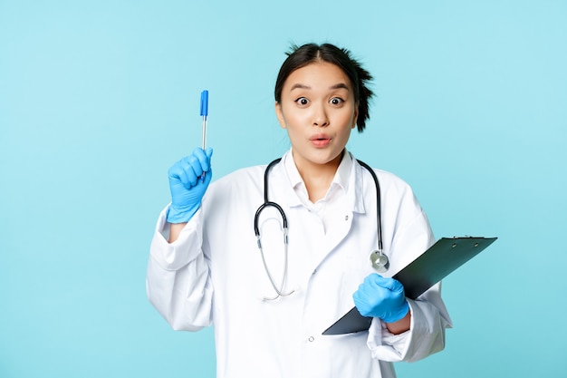 Excited asian female physician holding clipboard, raising pen up in eureka sign, has revelation, pitching an idea, standing on blue background