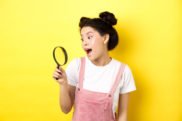 Photo excited asian girl found interesting thing, looking through magnifying glass amazed, standing on yellow summer