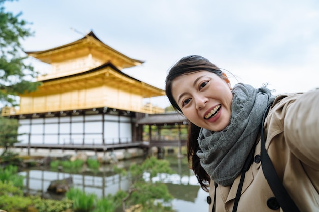 Excited asian girl is taking picture with the breathtaking
must-see in kyoto's heritage site. delightful asian visitor is
taking selfie with the gold leaf adorned kintakuji in japan on a
sunny day.