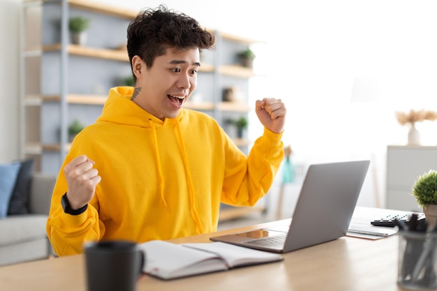 Excited asian guy using laptop celebrating success shaking fists