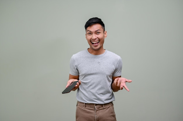 Excited Asian man holding his phone and opening his palm standing against an isolated background